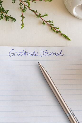 Op een witte tafel ligt een open spiraalvormig notitieboek met het handgeschreven opschrift "Gratitude Journal". Een zilveren pen rust op de pagina. Ernaast staat een kopje lichtgekleurde thee, een waxinelichtje in een witte keramieken houder en een paar delicate, witte takjes bloemen - dit alles samen creëert een kalme, ontspannen sfeer.