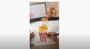 A hand refines the lettering "POP CORN" with a black pen in an almost finished hand lettering "Keep calm and eat more popcorn". The popcorn bag is watercoloured in red and yellow, next to it are Tombow pens and a glass of water.
