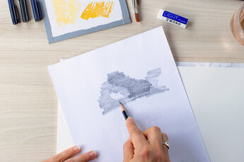 Une main hachure le verso d'un modèle de handlettering avec un crayon. Cette technique sert à reporter plus tard les lignes sur une nouvelle feuille en les traçant. En arrière-plan se trouvent divers ustensiles de dessin tels qu'une gomme, des pinceaux et des études de couleurs.