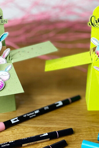 [Deux cartes de Pâques multicolores en forme de boîtes dépliées sont posées sur une table en bois. Chaque boîte est décorée de lapins de Pâques dessinés dans des couleurs vives et d'herbe en papier. Sur les deux cartes, on peut lire "Joyeuses Pâques". Au premier plan se trouvent des feutres roses, bleus et noirs.