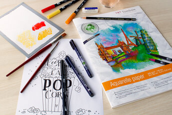Workstation with Tombow materials for creative hand lettering and watercolour: coloured Tombow ABT Dual Brush Pens, Tombow MONO fineliners, watercolour brushes, pencils and erasers next to a sketch sheet with popcorn hand lettering and a pad of watercolour paper.