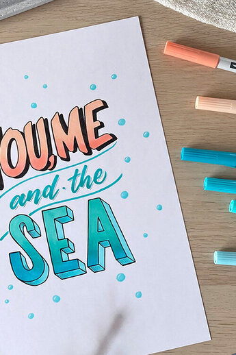 De afbeelding toont een zomers handschrift met de woorden "YOU, ME and the SEA" in koraal- en blauwtinten. Zes ABT PRO markers van Tombow in bijpassende kleuren liggen ernaast - perfect voor frisse, maritieme ontwerpen.