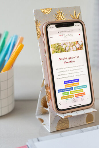 Le porte-téléphone DIY terminé est stable sur une table et tient un smartphone. Il est recouvert de papier ananas doré brillant. À côté, il y a des stylos colorés dans un gobelet et un carnet de notes en forme de cactus - idéal pour le bureau.