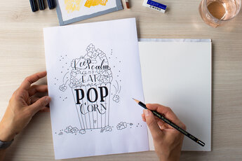 One hand holds a black and white template with hand lettering showing a popcorn bag with the words "Keep calm and eat more popcorn". Next to it is a blank drawing pad, ready for the drawing to be transferred. There are drawing materials in the background.
