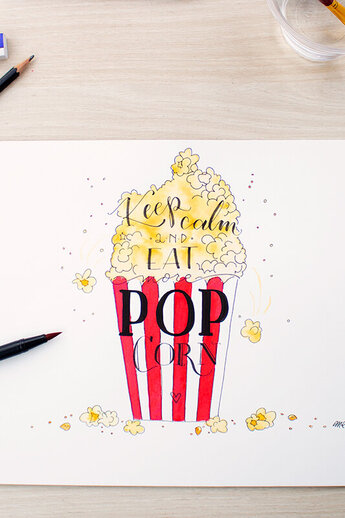 Handgemaakt handschrift met een rood-wit gestreepte popcornzak en de woorden "Keep calm and eat more popcorn", ingekleurd met Tombow ABT Dual Brush Pens. Omringd door een potlood, gum en verschillende Tombow pennen.