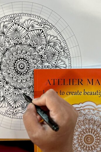 Een gedetailleerde mandala-tekening wordt uitgewerkt met een fineliner. Het precieze patroon volgt een geometrische rasterstructuur die symmetrische vormen en ornamenten ondersteunt. Op de voorgrond staat het boek "ATELIER MANDALA - Leer prachtige mandala's maken", dat dient als inspiratie of instructie.