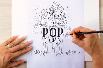 One hand traces details on a hand lettering template "Keep calm and eat more popcorn" while the other fixes the paper. The popcorn bag and lettering are already finished in black.