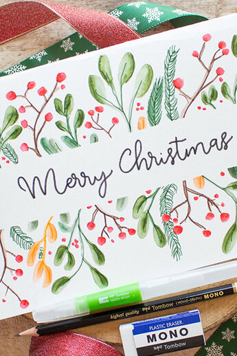 Kerstkaart met "Vrolijk kerstfeest" in kalligrafie, omringd door waterverfmaretak en rode bessen. Versierd met groene en rode strikken en gouden accenten. Tombow brush pennen, gum en water brush ernaast.