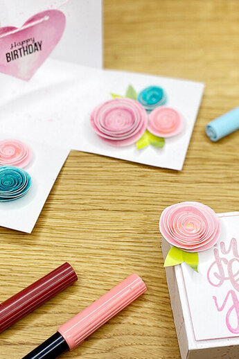 Een zelfgemaakte verjaardagskaart en cadeauverpakking zijn versierd met kleurrijke, handgemaakte papieren bloemen en letters. Rondom liggen borstelpennen en knutselmaterialen op een houten tafel.