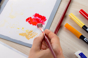 A hand picks up red paint from a mixing palette with a flat brush. Next to it are other colours, Tombow pencils in shades of yellow, orange and red and an eraser.