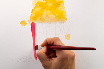 A hand paints a red stripe on the pre-drawn popcorn bag with a flat brush. The upper area with popcorn is already coloured in yellow, the pencil lines of the hand lettering "Keep calm and eat more popcorn" are still visible.