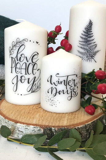 Handbeschriftete Kerzen mit festlichen Motiven und Schriftzügen wie „Love, Peace, Joy“ und „Winter Days“ stehen dekorativ auf einer Holzscheibe. Die Designs wurden mit Tombow MONO graph Bleistift skizziert und anschließend übertragen.