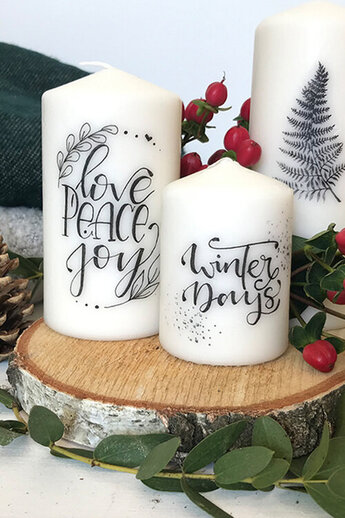 Met de hand ingegraveerde kaarsen met feestelijke motieven en letters zoals "Love, Peace, Joy" en "Winter Days" staan decoratief op een houten schijf. De ontwerpen zijn geschetst met Tombow MONO grafiekpotlood en daarna overgebracht.
