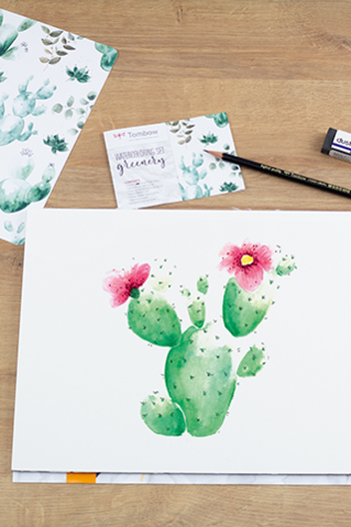 Een aquarel kunstwerk met een bloeiende cactus ligt omringd door tekenmaterialen op een houten tafel. Naast verschillende groene ABT Dual Brush Pens is er een watertankpenseel, een waterproof potlood, een Dust Catch gum en een mengpalet. Inspiratiekaarten met cactusmotieven en verpakkingen met het opschrift "Aquarelset Greenery" maken de creatieve opstelling compleet.